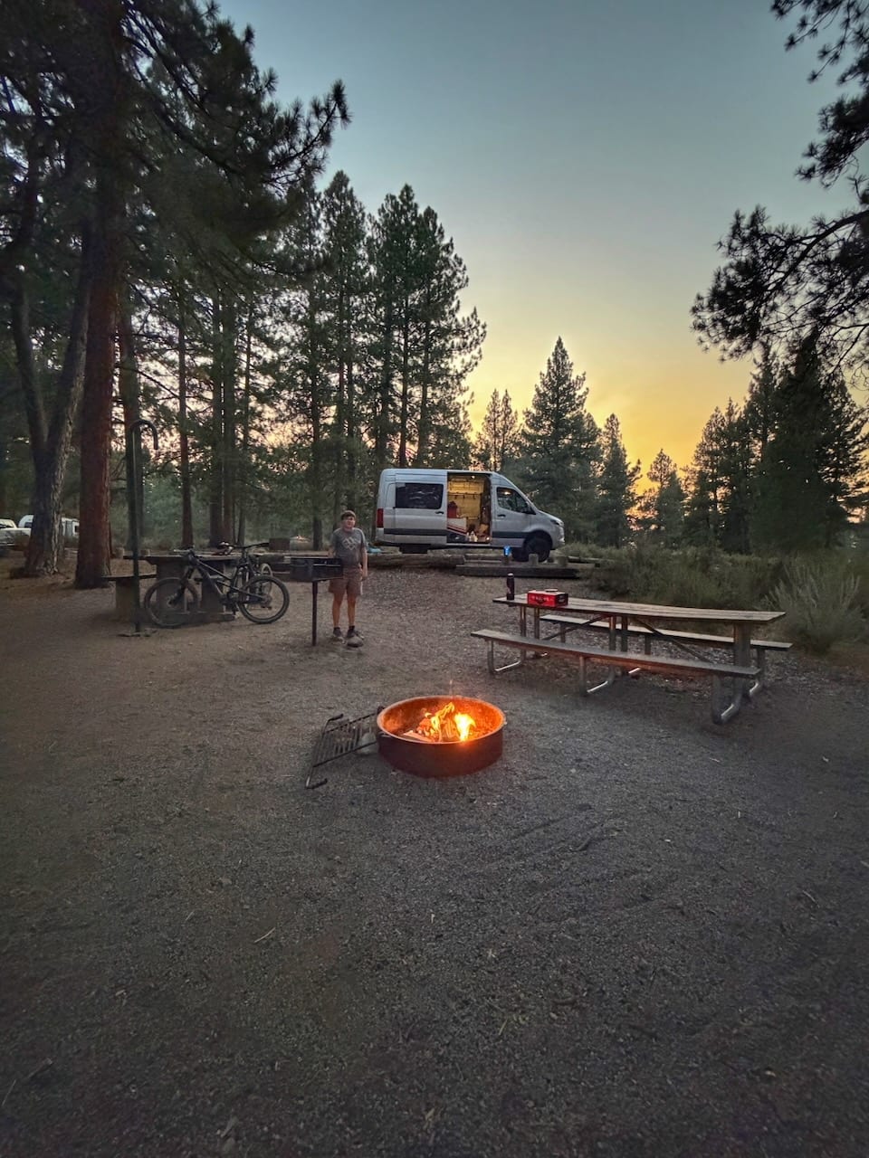 A campfire and picnic tables at a forest campsite at dusk, with the van parked behind