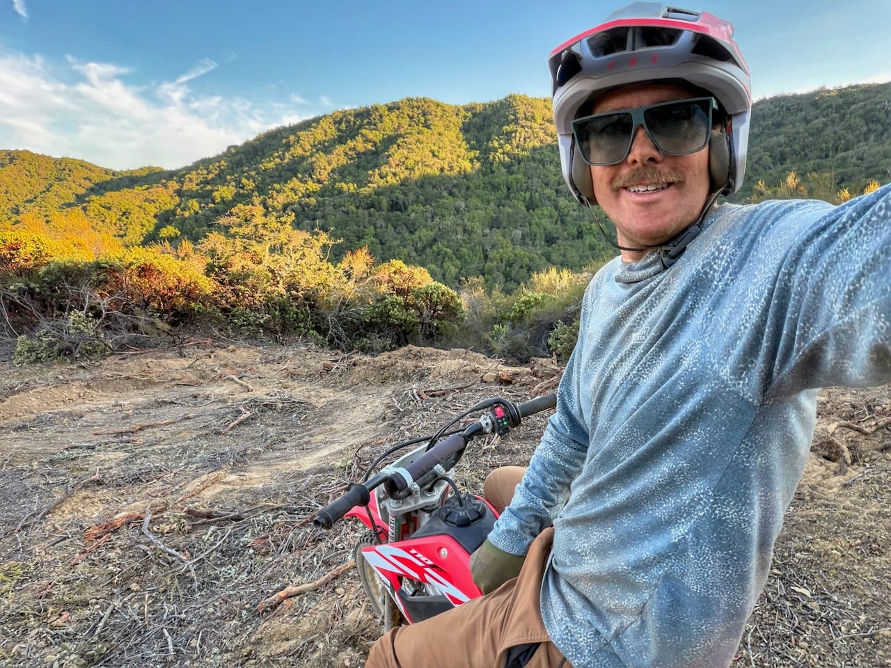 Jed in a helmet beside a dirt bike on a ridge above green hills