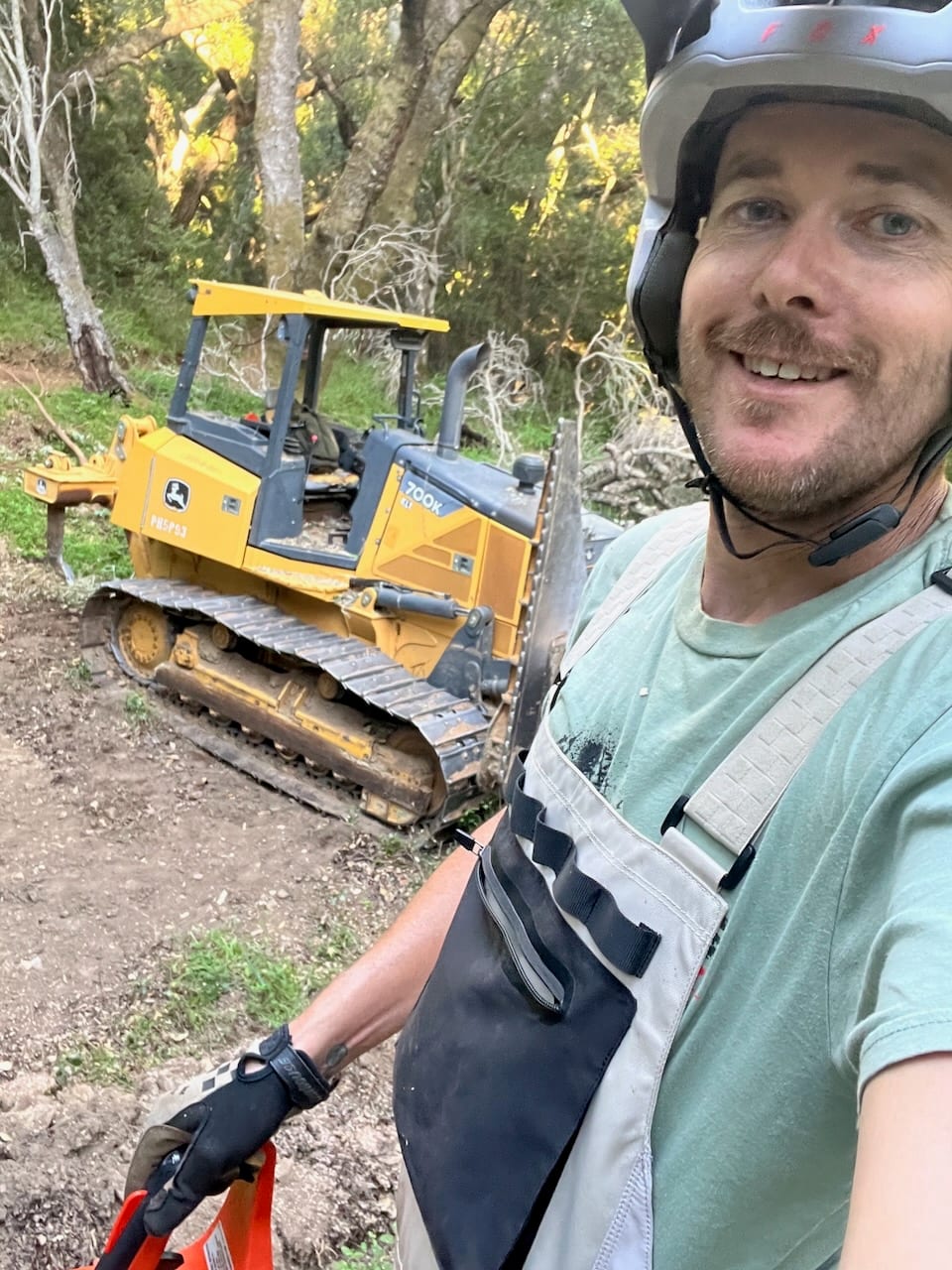 Jed in a helmet and work clothes in front of a small dozer on a new trail