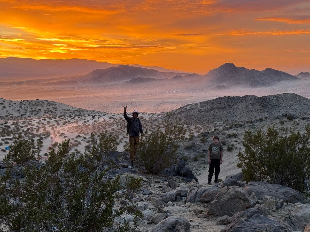 An adult in a helmet raising one hand and a boy standing on a rocky desert ridge under an orange sunset, with a dusty valley and mountains behind them
