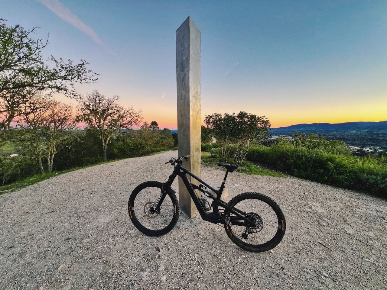 A mountain bike leaning against a tall metal pillar on a gravel hilltop at sunset, with a valley below