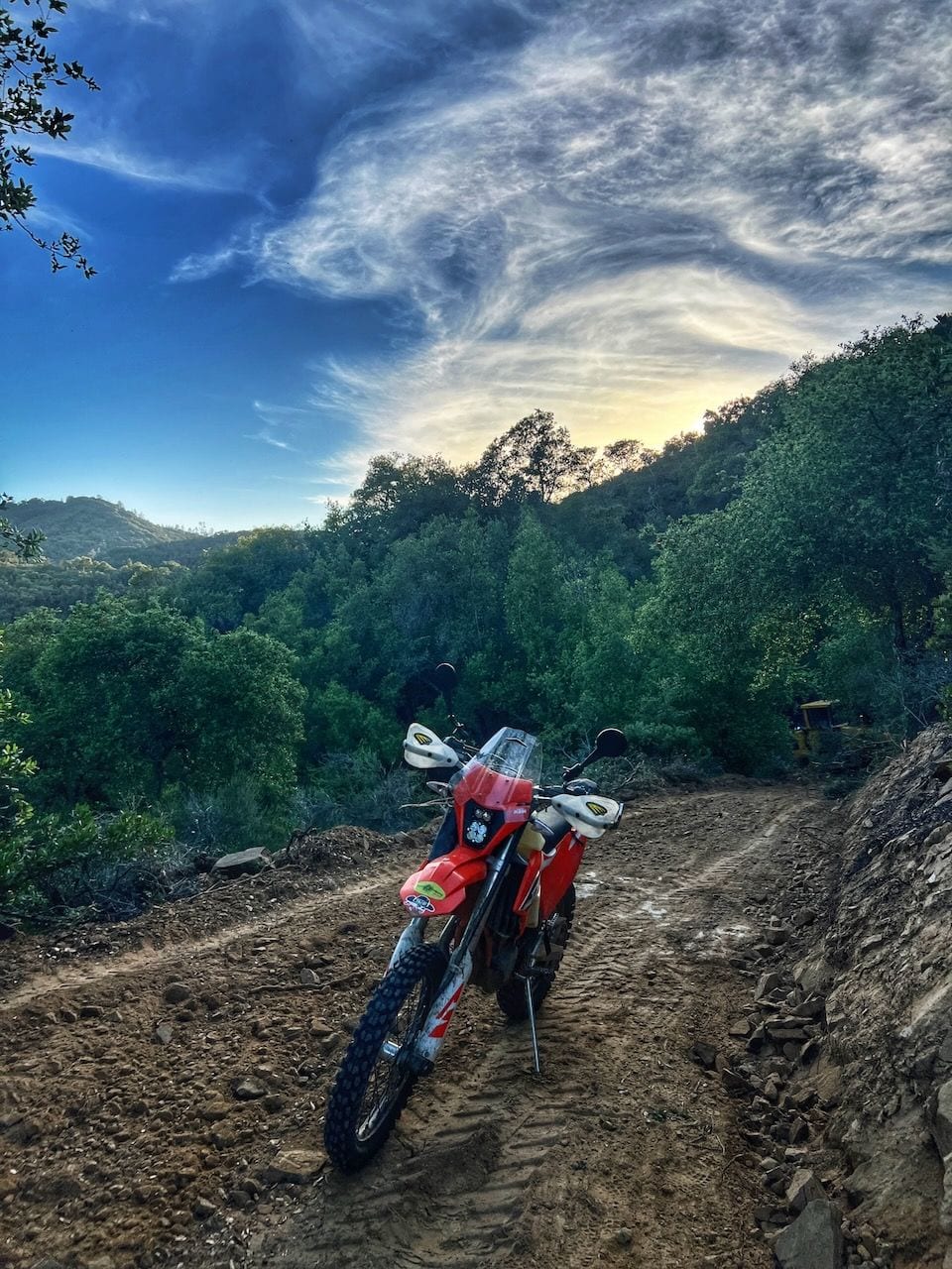 A red dirt bike on a freshly cut dirt road through wooded hills, with yellow equipment parked in the distance