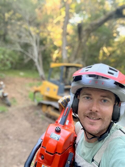 Jed in a helmet and ear protection holding a chainsaw on his shoulder, with a small dozer behind him