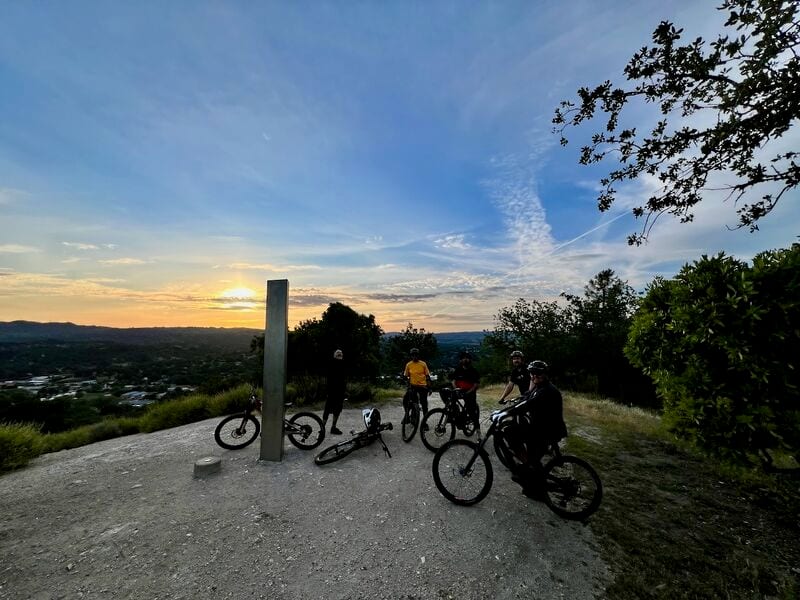 A group of mountain bikers with their bikes on a gravel hilltop at sunset, next to a tall metal post