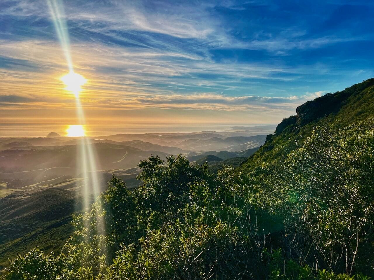 The sun setting over the ocean, seen from a brushy ridge above rolling coastal hills