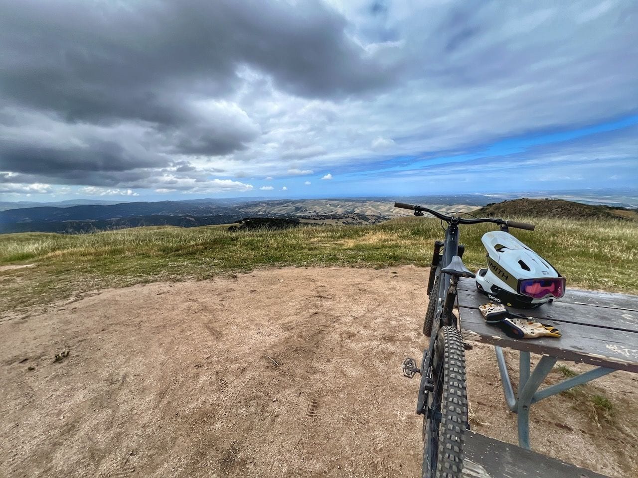 A mountain bike leaning on a picnic table with a helmet and gloves, on a grassy hilltop under a cloudy sky