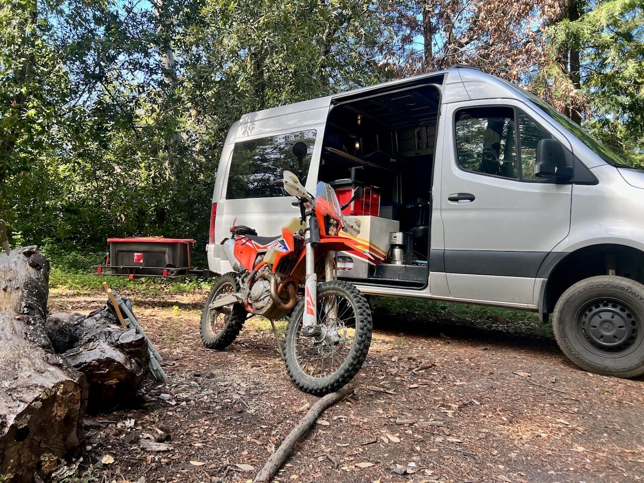 An orange dirt bike parked beside a silver camper van with its side door open at a wooded campsite