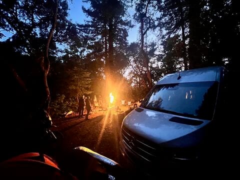 A campfire glowing between tall trees at dusk, with people standing around it and a camper van parked nearby
