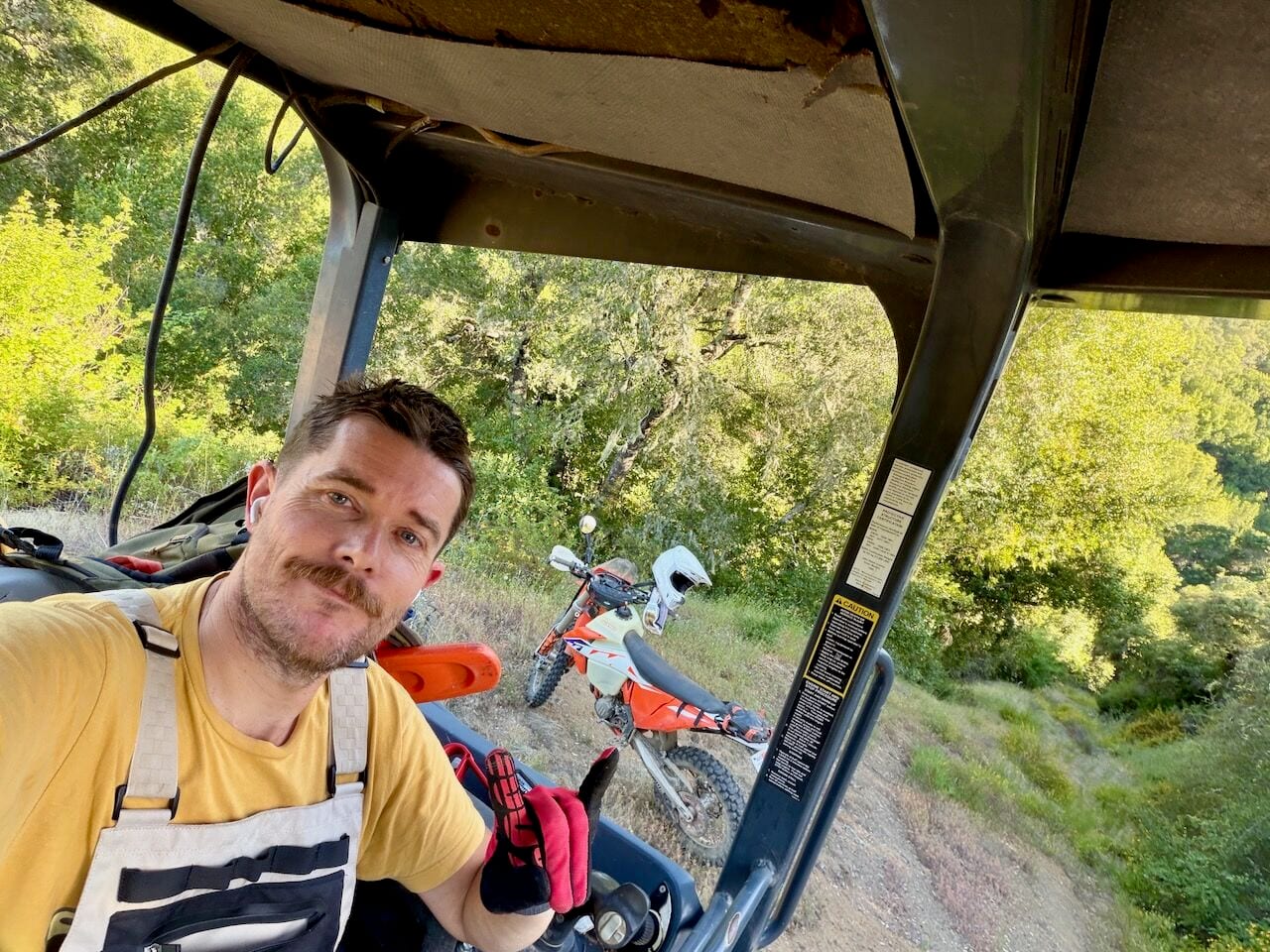 Jed in a yellow shirt and riding gear taking a selfie from a side-by-side, with an orange dirt bike parked on a wooded trail behind him