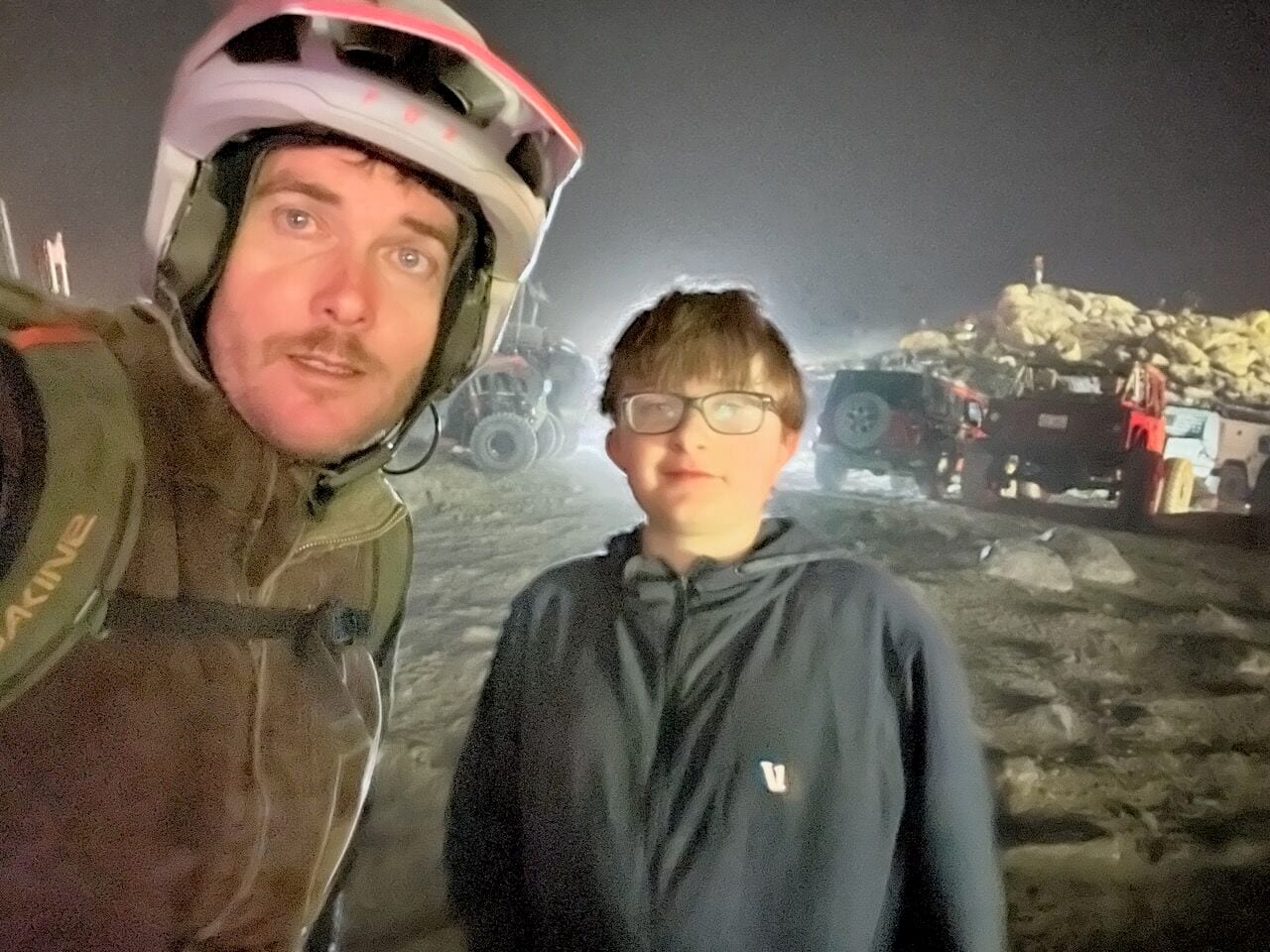 Jed in a helmet and his son in a hooded jacket in a night selfie, with off-road vehicles under bright lights behind them