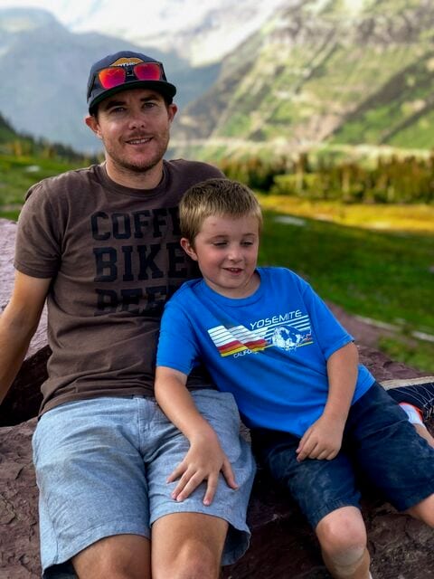 A man and a young boy in a Yosemite T-shirt sitting on a rock in a mountain meadow